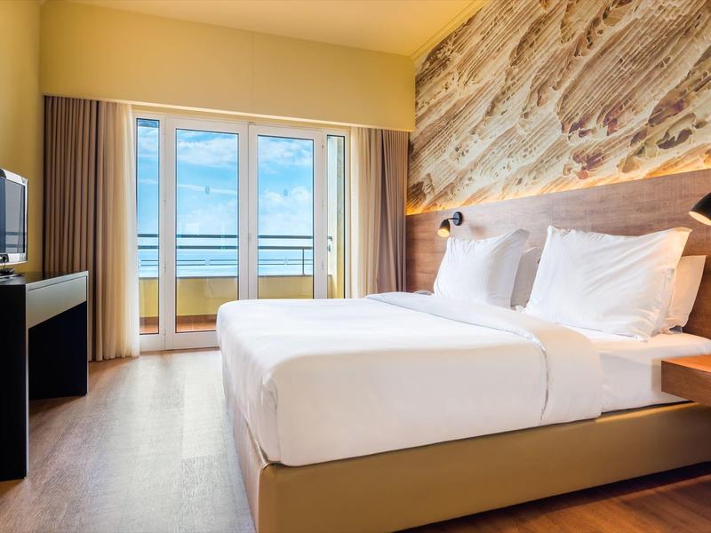 Helles Schlafzimmer mit Doppelbett, Fensterfront, Meerblick und moderner Wandgestaltung aus Stein.