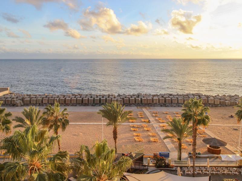 Strand mit Palmen und Liegestühlen, darüber bewölkter Himmel und ruhiges Meer im Sonnenuntergang.