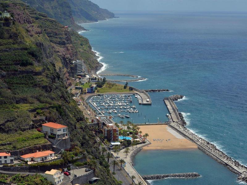 Küstenlandschaft mit Hafen, Sandstrand, grünen Hügeln und blauem Meer unter bewölktem Himmel.