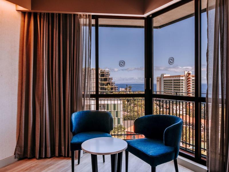 Hotelový pokoj se dvěma modrými křesly a výhledem na město přes velká okna.