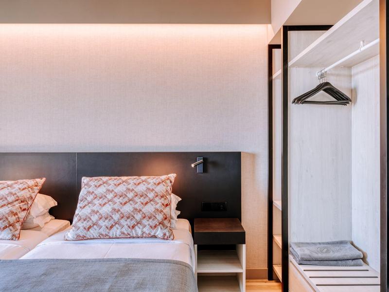 Moderní hotelový pokoj se dvěma postelemi, nočním stolkem a otevřenou skříní.