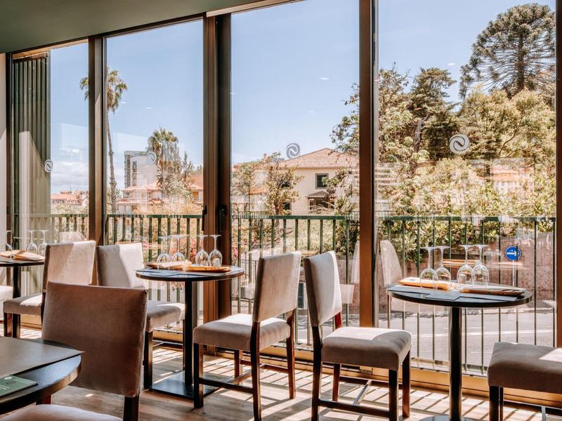 Příjemná snídaňová část s stoly a židlemi u velkých skleněných oken s výhledem na stromy a budovy.