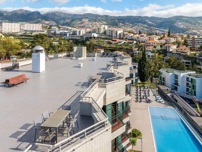 Střešní terasa s lehátky s výhledem na bazén a město s horami v pozadí.