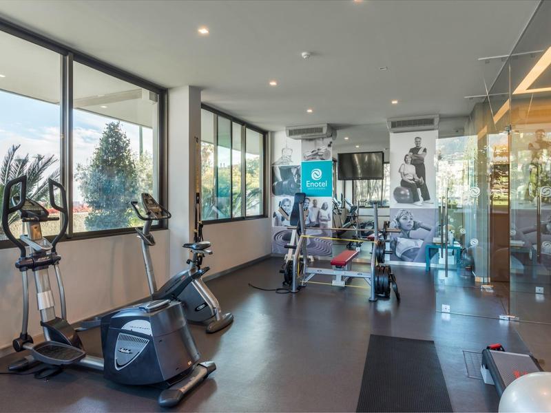 Moderní posilovna s cvičebními stroji a velkými okny s výhledem do zeleně.