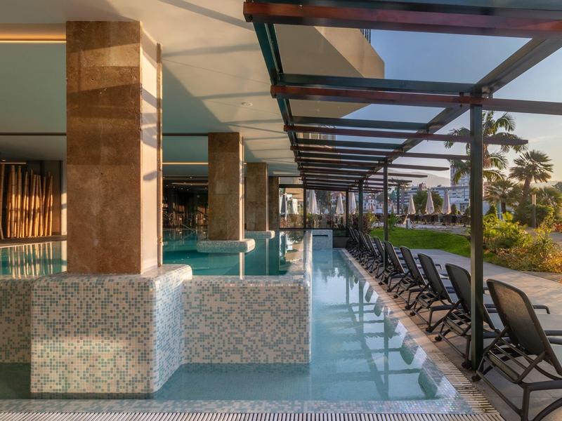 Moderní hotelová bazénová plocha s lehátky a přístřeškem vedle bílé budovy.