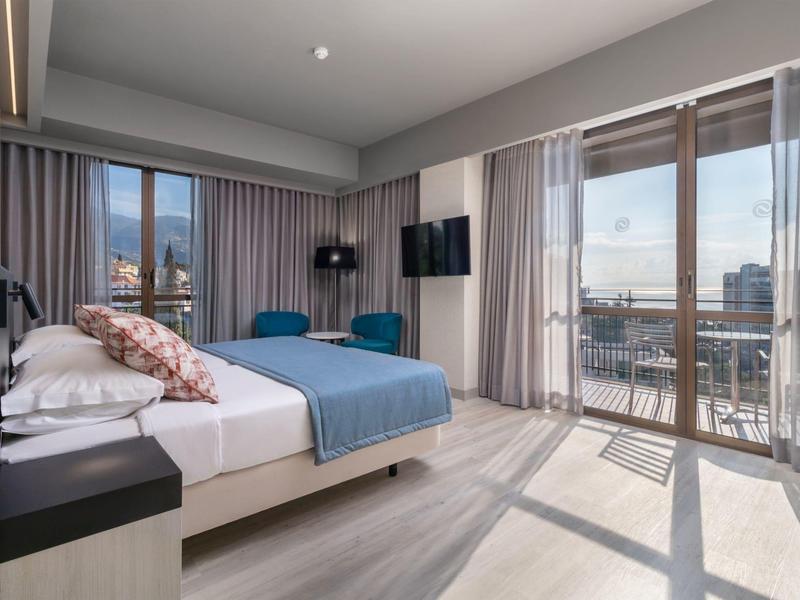 Světý hotelový pokoj s manželskou postelí, modrou pohovkou a balkonem s výhledem na město.