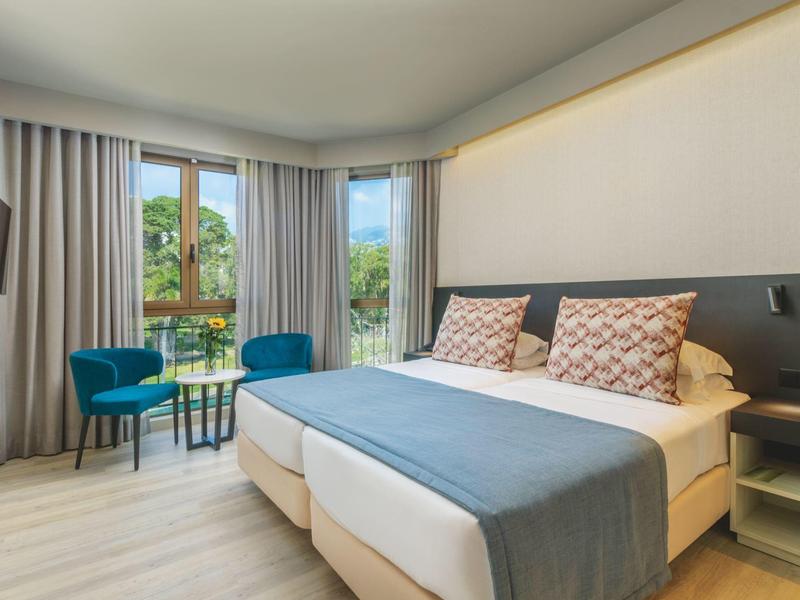 Jasně zařízený hotelový pokoj se dvěma postelemi, modrými židlemi a přístupem na balkon.