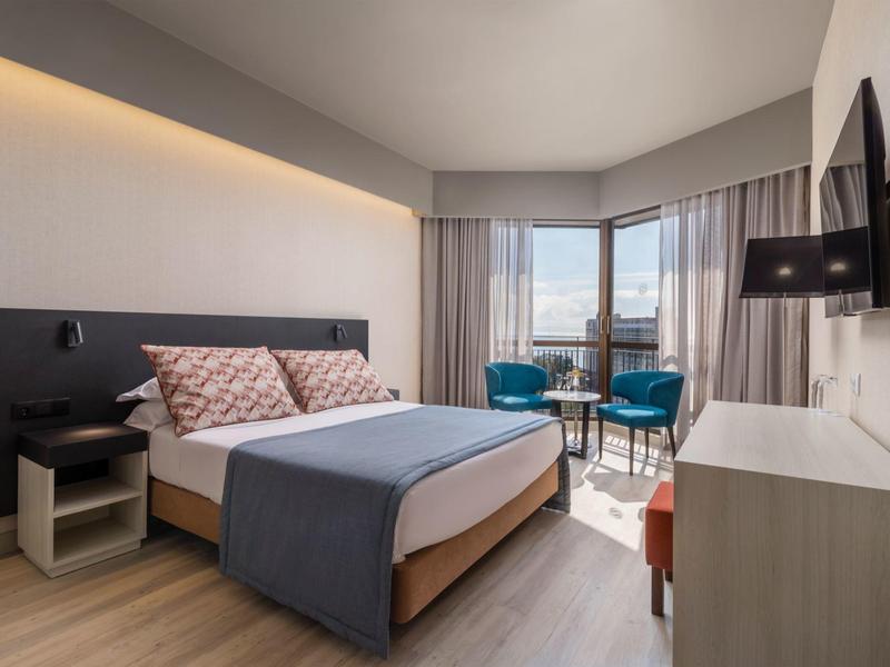 Moderní hotelový pokoj s velkou postelí, křesly u okna a televizí na stěně.