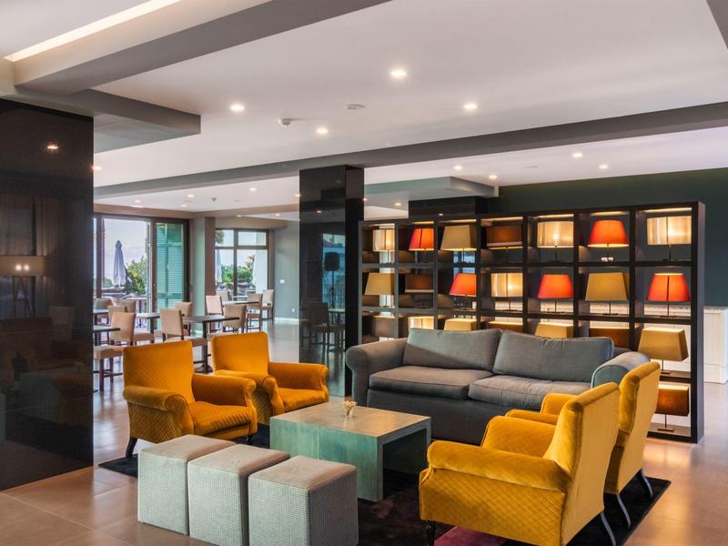 Moderní hotelová hala se šedou pohovkou, oranžovými křesly a dekorativními regály s lampami.