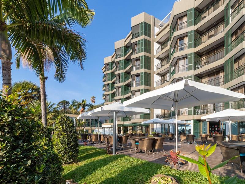 Venkovní prostor hotelu s slunečníky, palmami a vícepatrovou budovou v pozadí.