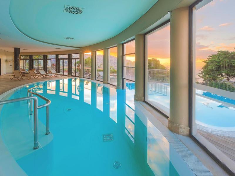 Vnitřní bazén s velkými okny s výhledem na venkovní bazén při západu slunce.