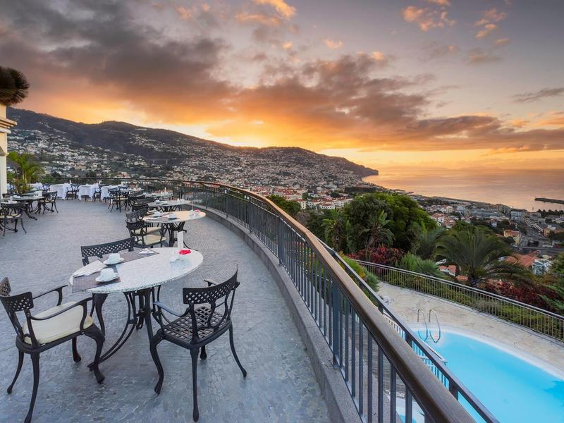 Terasa s stolem a židlemi, výhled na město na kopci a západ slunce nad mořem