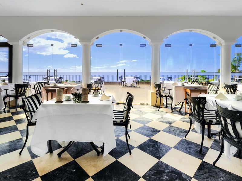 Restaurace s výhledem na moře a černobílou podlahou pod bílými oblouky.