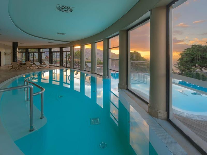 Vnitřní bazén s velkými okny s výhledem na venkovní bazén a západ slunce.