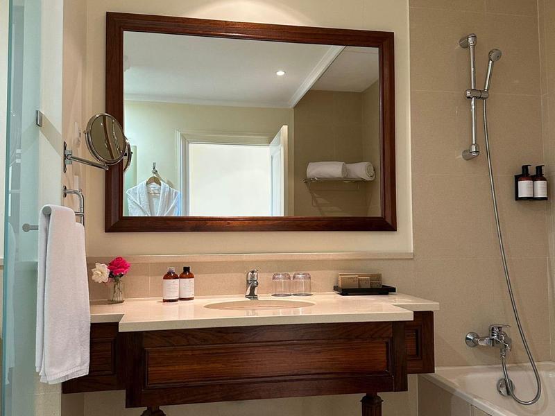 Jemně osvětlená koupelna se dřevěným nábytkem, velkým zrcadlem a vanou vedle umyvadla.