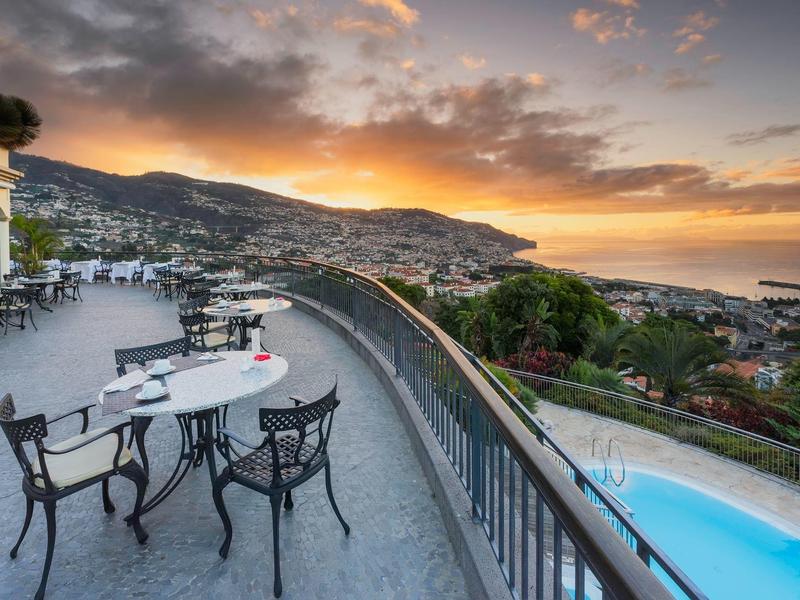 Terasa s lavičkami a židlemi s výhledem na město, hory a moře při západu slunce