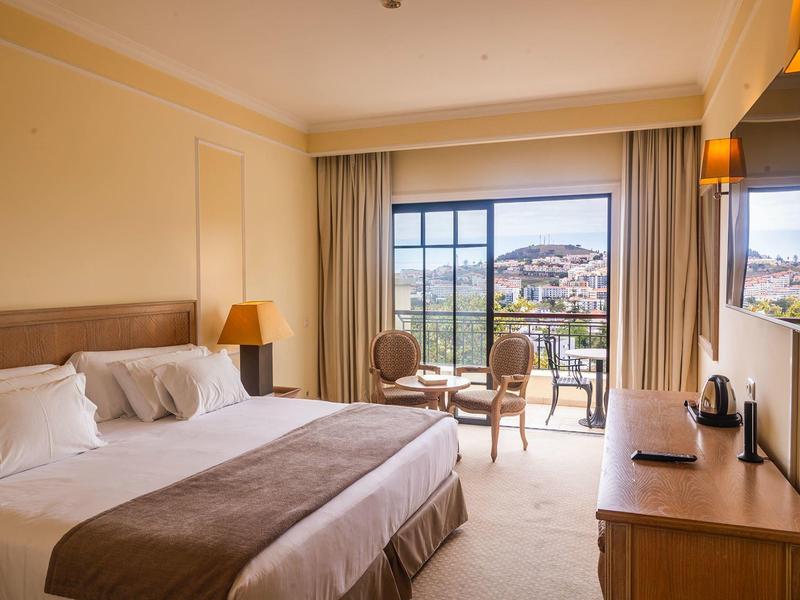 Světý hotelový pokoj s manželskou postelí, posezením a balkonem s výhledem na město.