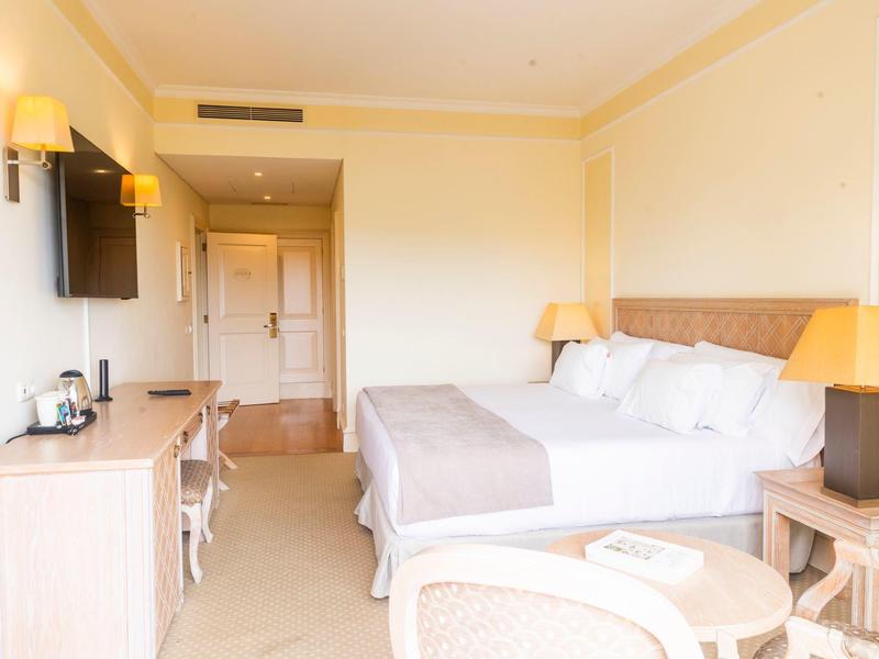 Světý hotelový pokoj s manželskou postelí, psacím stolem, televizí a posezením.