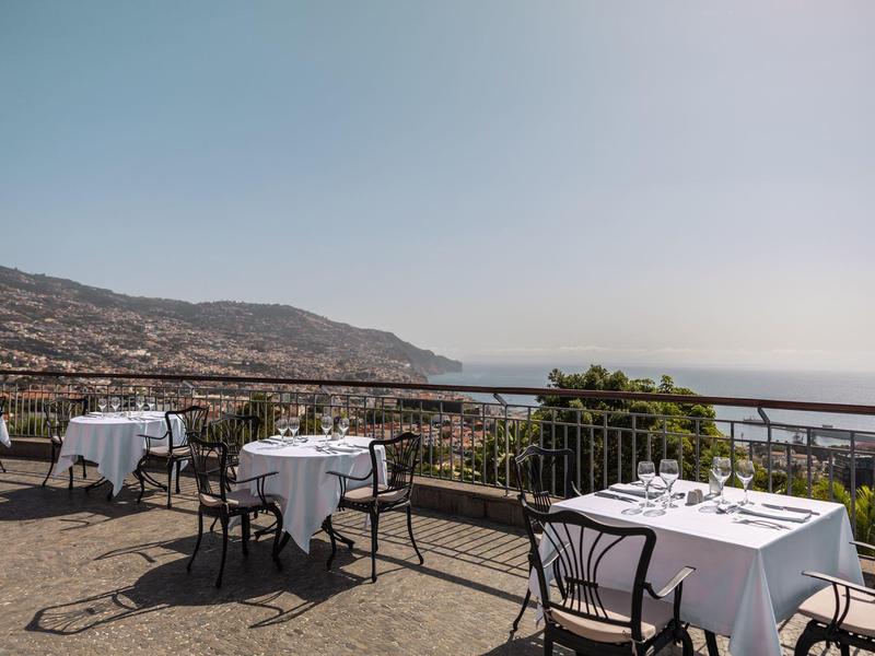 Terasa s prostřenými stoly, výhled na město a moře za jasné oblohy.