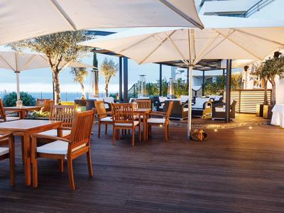 Terrasse mit Holzboden, weißen Sonnenschirmen, Holztischen und Stühlen mit Blick auf das Meer bei Sonnenuntergang