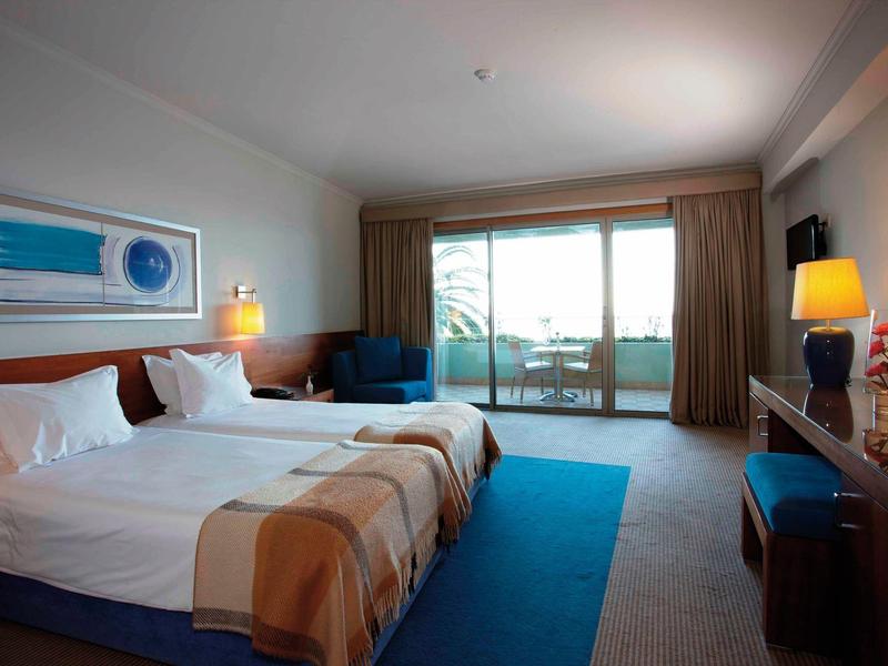 Hotelzimmer mit Doppelbett, blauen Teppichen, Sitzbereich und Balkon mit Blick nach draußen.