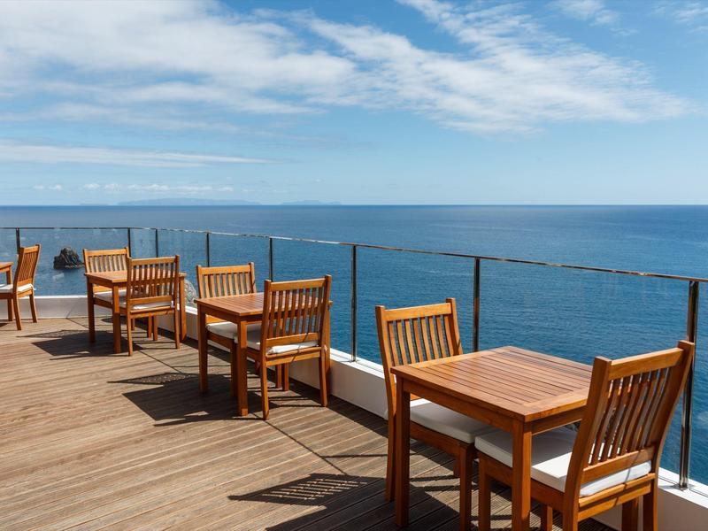 Terrasse mit Holzboden und Holzstühlen mit Tischen mit Blick aufs ruhige Meer unter blauem Himmel.