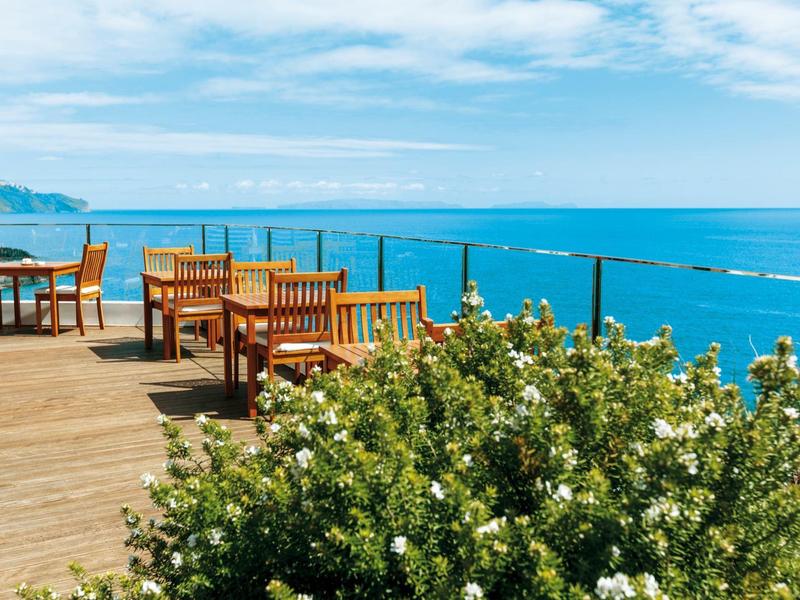 Terrasse mit Holztischen und Stühlen mit Blick auf das blaue Meer und bewölkten Himmel