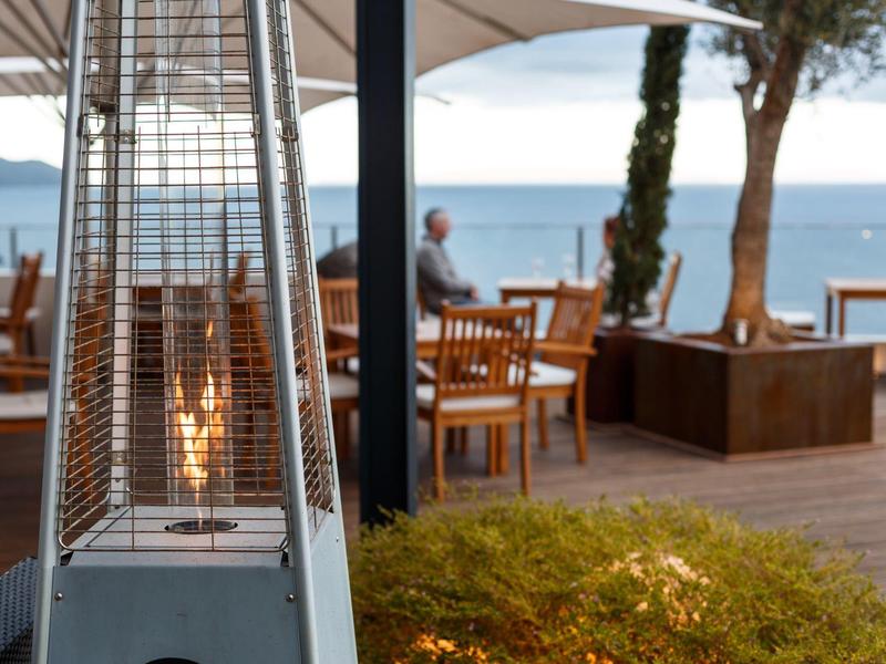 Terrasse mit Holzstühlen und Stehelement mit Flamme, Blick aufs Meer bei Sonnenuntergang.
