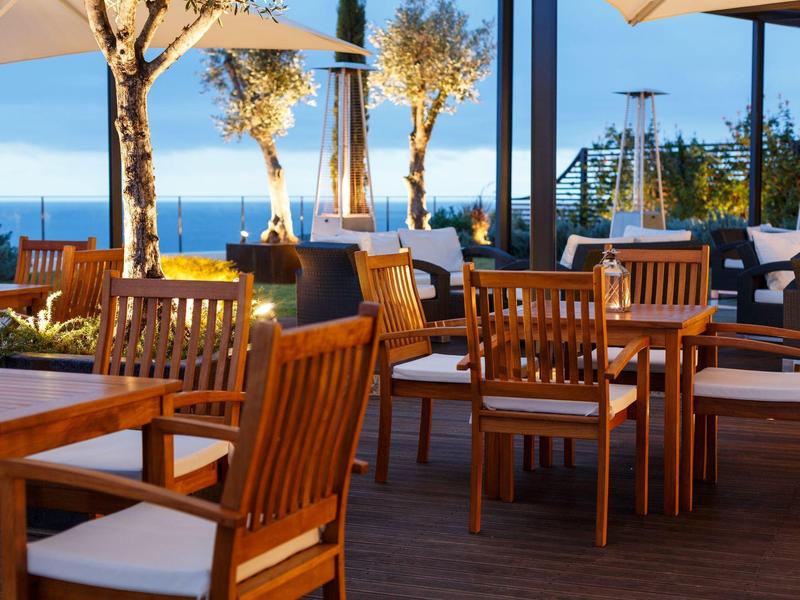 Außenrestaurant mit Holztischen und Stühlen auf einer Terrasse mit Meerblick bei Sonnenuntergang.