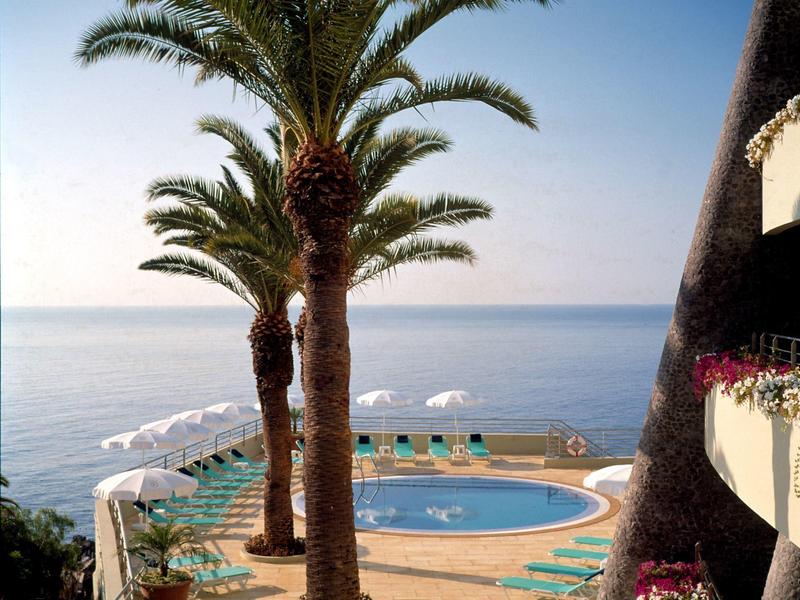 Poolbereich mit Liegestühlen und Palmen mit Blick auf das Meer an einem sonnigen Tag.