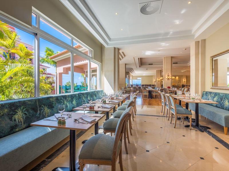 Ristorante luminoso con tavoli moderni, sedie e grandi finestre con vista su piante tropicali.