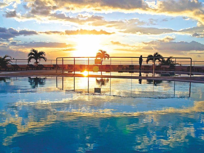 Complesso alberghiero con piscina vicino al mare, circondato da alberi e cielo azzurro.
