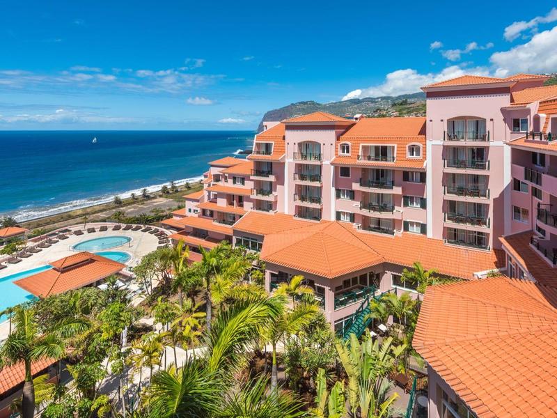 Complesso alberghiero con piscina, palme e vista sul mare sotto un cielo blu.
