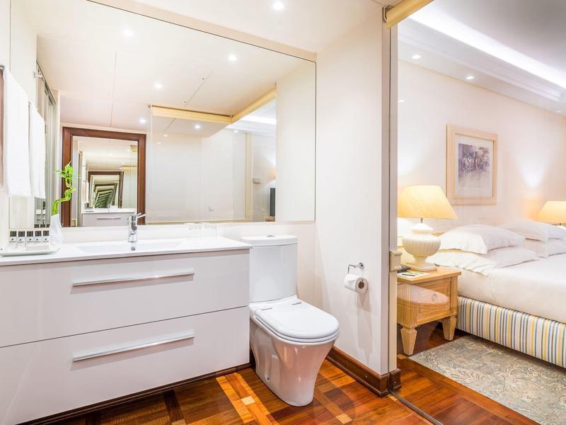 Bagno moderno con grande specchio, mobili bianchi e camera da letto adiacente.