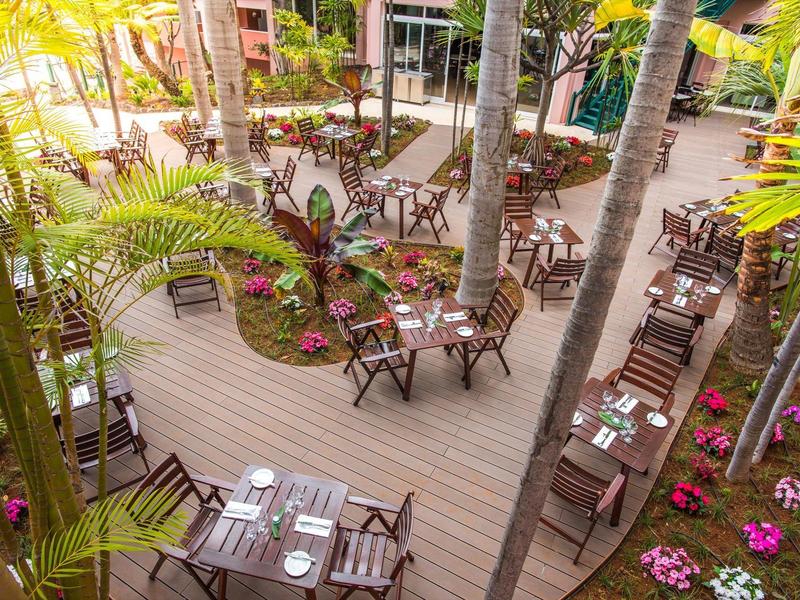 Accogliente area esterna dell'hotel con tavoli, sedie e palme tra aiuole fiorite.