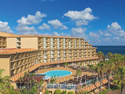 Hotel de varios pisos con piscina redonda y palmeras bajo un cielo azul junto al mar.