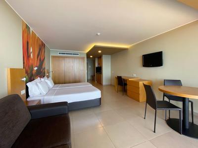 Habitación de hotel moderna con cama doble, televisión, mesa, sillas y sofá en tonos neutrales.