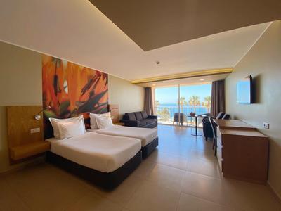 Habitación de hotel moderna con dos camas, zona de estar y vista al mar a través de grandes ventanas.