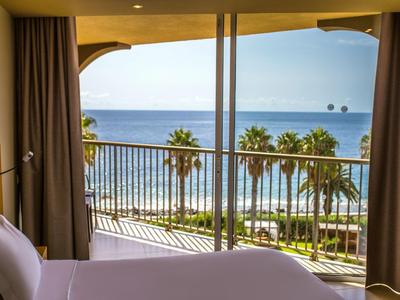 Vista desde la habitación del hotel con balcón al mar y palmeras bajo cielo claro.