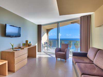 Habitación de hotel moderna con sofá, sillón, mesa, TV y vista al mar desde el balcón.