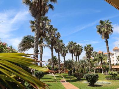 Jardín verde con palmeras y caminos frente a un edificio de hotel bajo un cielo azul.