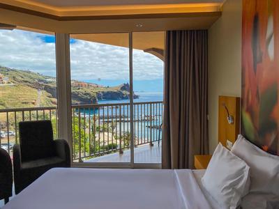 Habitación de hotel con cama grande, ventana panorámica y vista al mar y montañas.