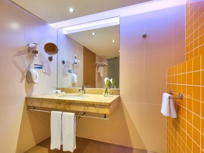 Baño moderno de hotel con espejo grande, lavabo y azulejos naranjas en la pared.