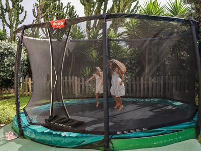 Niños saltando en una cama elástica redonda en el jardín, rodeados de árboles y arbustos.