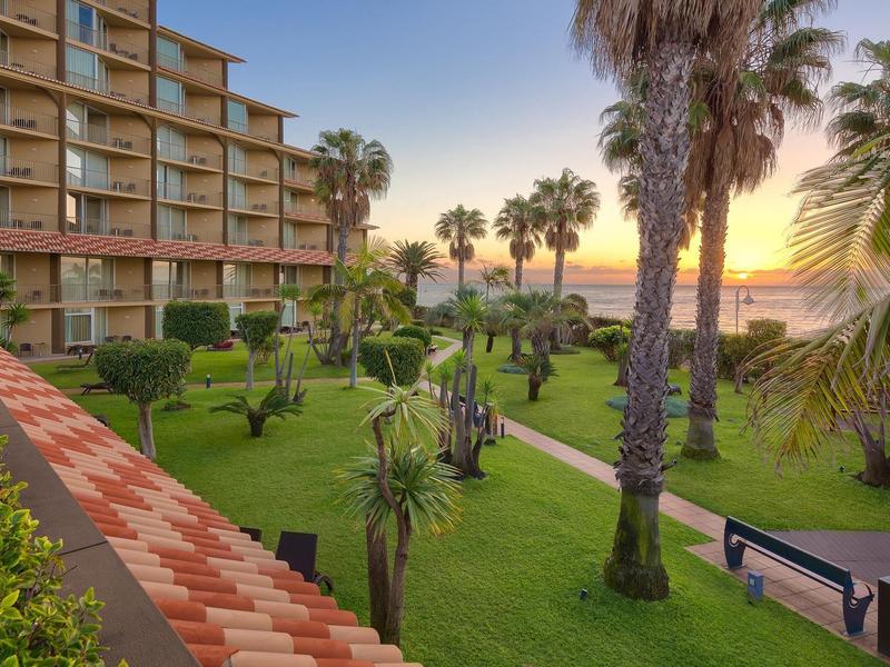 Edificio de hotel con jardín, palmeras y bancos al atardecer junto al mar