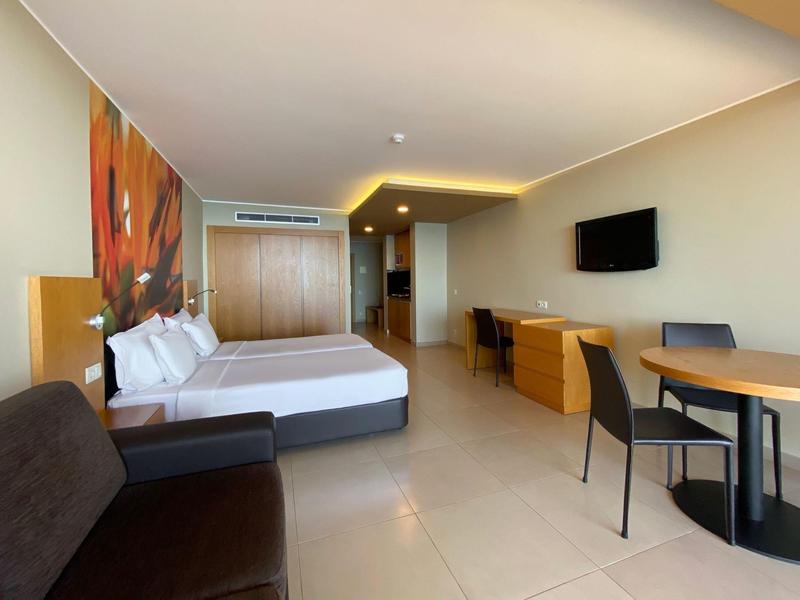 Habitación de hotel moderna con cama doble, televisión, mesa, sillas y sofá en tonos neutrales.