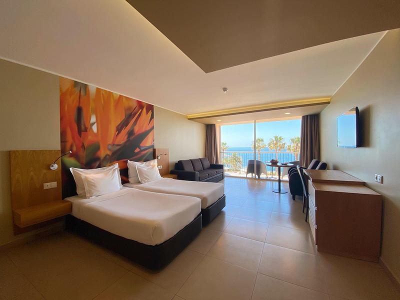 Habitación de hotel moderna con dos camas, zona de estar y vista al mar a través de grandes ventanas.