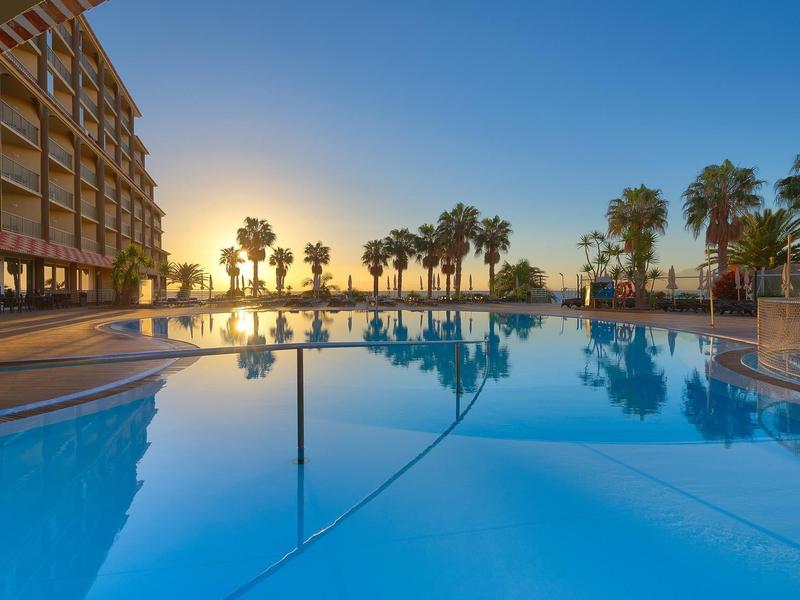Piscina del hotel con palmeras al atardecer y agua clara que refleja el entorno.