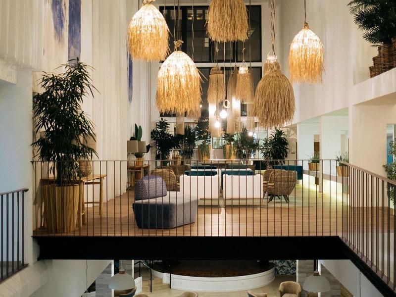 Interior moderno de hotel con varias áreas de asiento, grandes plantas y lámparas colgantes llamativas.