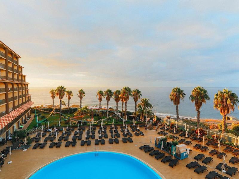 Terraza de hotel con piscina, tumbonas y palmeras con vistas al mar al atardecer.